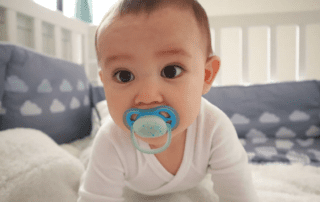 Baby in white onesie with pacifier in mouth.
