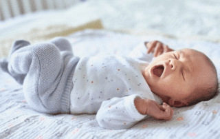 Newborn yawing, lying on a bed in a white and grey onesie.