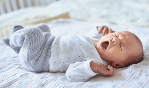 Newborn yawing, lying on a bed in a white and grey onesie.